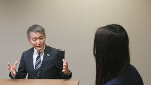 Man in a dark suit speaks and gestures while seated at a wooden table, facing a woman with long dark hair in an office setting.
