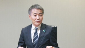 Middle-aged man in a dark suit and striped tie seated at a table, speaking thoughtfully in a meeting room
