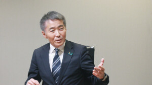 Middle-aged man in a dark suit and striped tie speaks while gesturing with his hand during a meeting.