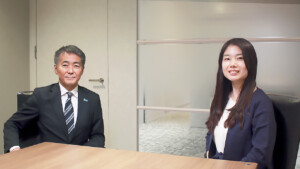 Two colleagues seated at a conference table in an office, smiling at the camera.