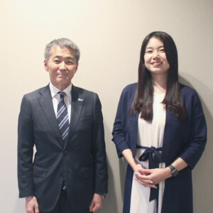 Two professionals posing for a photo: a gray-haired man in a suit and a young woman in a navy blazer, smiling against a plain wall.