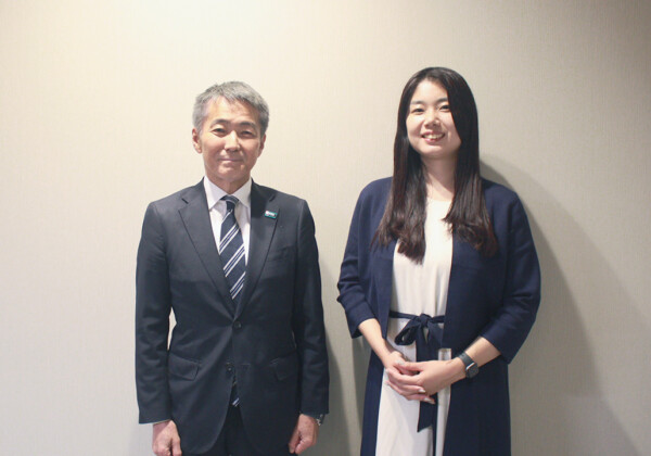 Two professionals posing for a photo: a gray-haired man in a suit and a young woman in a navy blazer, smiling against a plain wall.