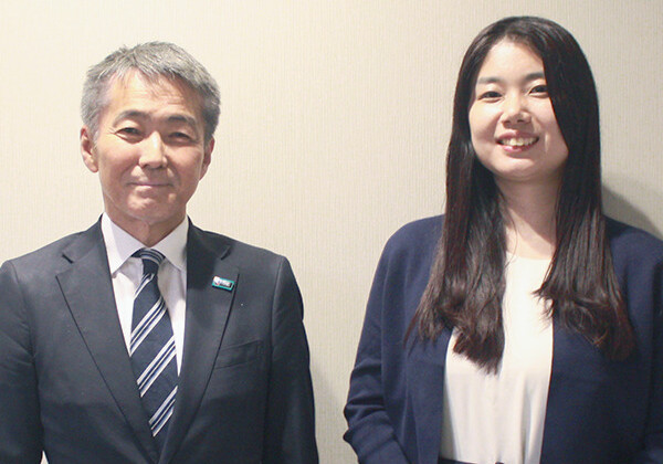 Man in a dark suit and striped tie stands beside a smiling woman in a navy blazer against a plain wall, posing for a photo in an office setting.