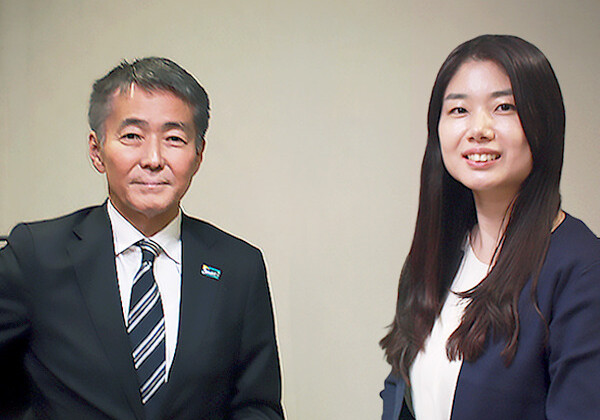 Two professionals sit at a desk in a neutral room, a man in a suit and striped tie beside a woman in a navy blazer smiling at the camera.