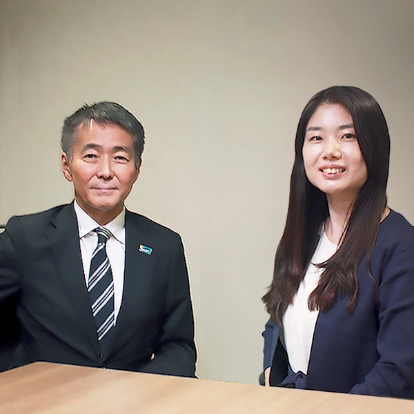 Two professionals sit at a desk in a neutral room, a man in a suit and striped tie beside a woman in a navy blazer smiling at the camera.