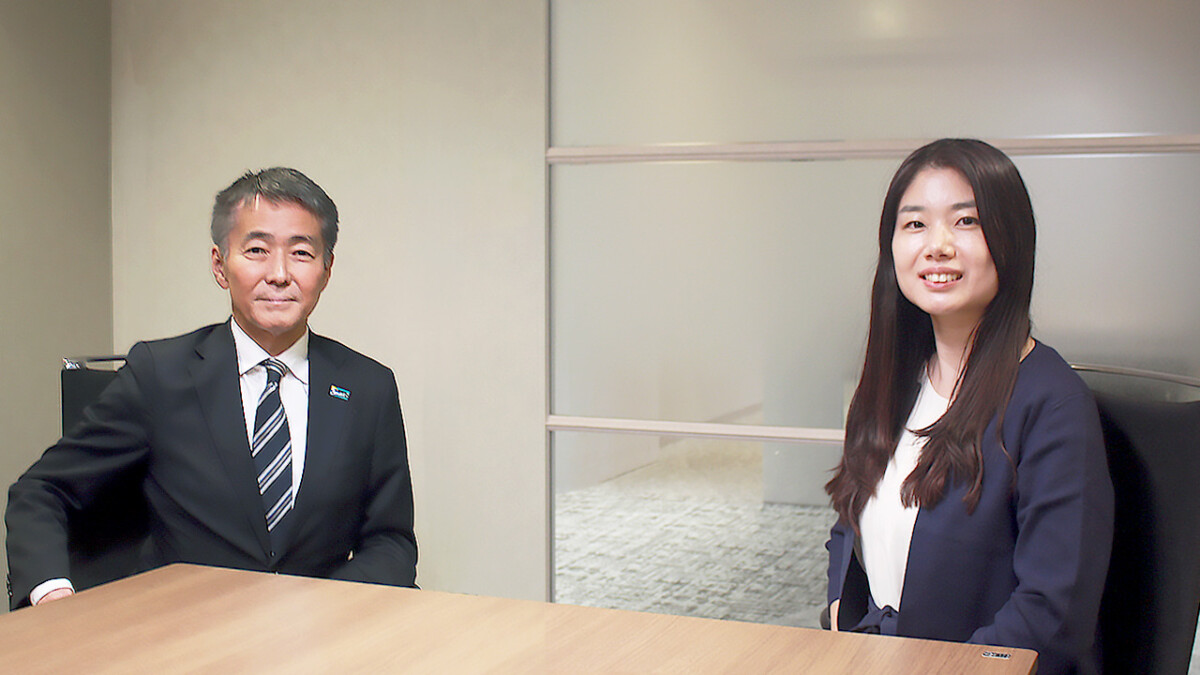 Two business professionals sit at a wooden conference table, smiling at the camera in a modern office setting.
