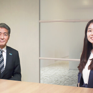 Two business professionals sit at a wooden conference table, smiling at the camera in a modern office setting.