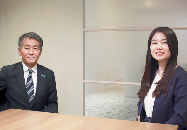 Two business professionals sit at a wooden conference table, smiling at the camera in a modern office setting.