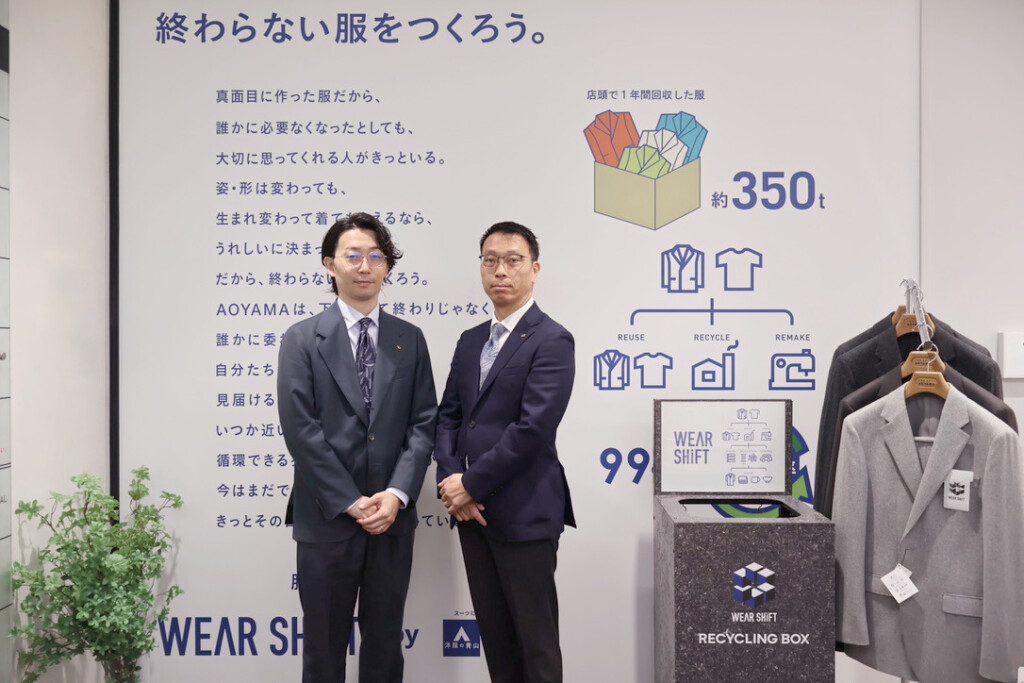 Two men in dark suits pose in front of a clothing recycling display with a box of folded clothes and garment icons on the wall.