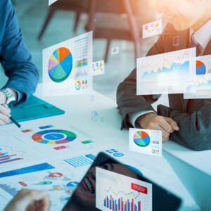 Two professionals in suits review colorful charts and graphs on a table during a meeting in a glass-walled office scenario.