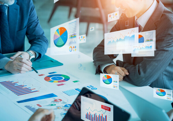 Two professionals in suits review colorful charts and graphs on a table during a meeting in a glass-walled office scenario.