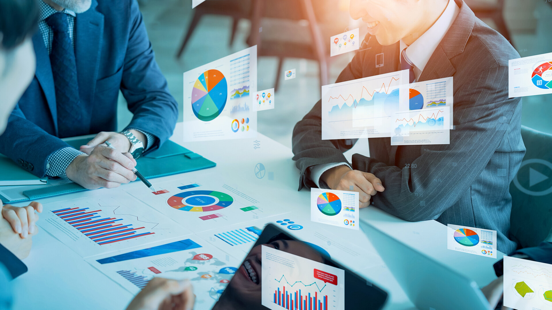 Two professionals in suits review colorful charts and graphs on a table during a meeting in a glass-walled office scenario.