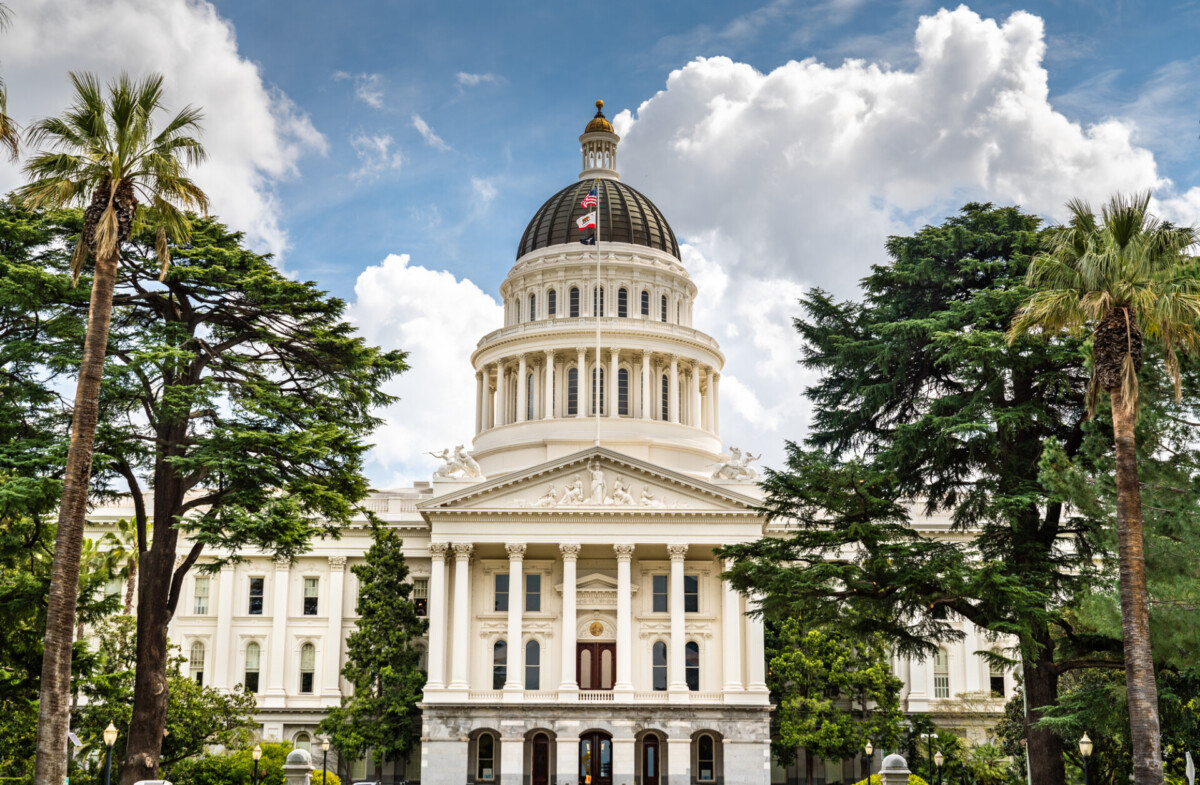Front view of a neoclassical capitol building with a large dome, flags, and surrounding palm trees under a blue sky.